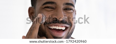 Close up head shot portrait overjoyed African American handsome young man applying moisturizing face cream, facial massage, enjoying skincare procedure, looking at camera, wide cropped image