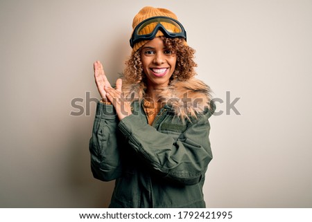 Young african american skier woman with curly hair wearing snow sportswear and ski goggles clapping and applauding happy and joyful, smiling proud hands together