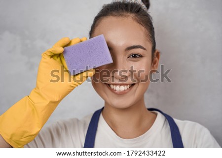 Close up portrait of funny cleaning lady in yellow rubber gloves covering one eye with kitchen sponge, looking at camera and smiling, standing against grey wall