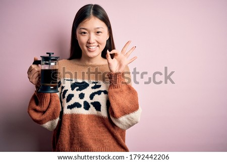 Young asian woman making a glass of coffe using french press coffee maker over pink background doing ok sign with fingers, excellent symbol