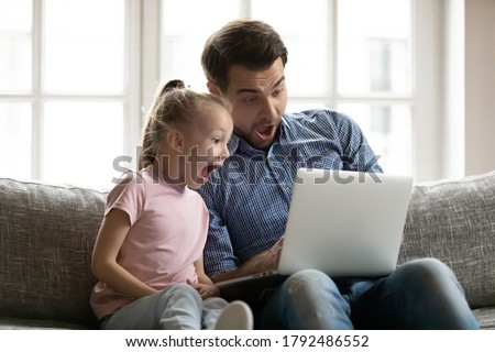 Emotional little cute child daughter watching funny movie on computer with joyful dad, sitting together on couch. Surprised two generations family looking at laptop screen, shocked by sale offer.