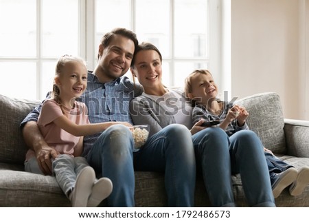 Happy bonding family of four resting on comfortable sofa, watching movie together indoors. Smiling young parents cuddling little children siblings, enjoying funny TV series with popcorn at home. Royalty-Free Stock Photo #1792486573