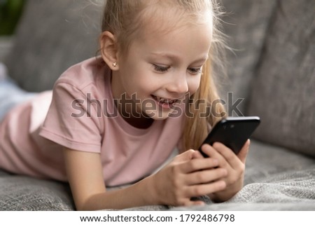 Close up smiling little cute child girl lying on comfortable sofa, involved in online mobile game. Addicted to technology happy adorable laughing blonde kid watching funny video in social networks.