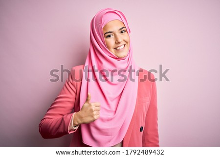 Young beautiful girl wearing muslim hijab standing over isolated pink background doing happy thumbs up gesture with hand. Approving expression looking at the camera showing success.