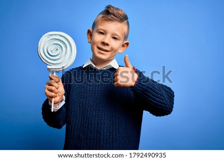 Young little caucasian kid eating sweet candy lollipop over blue isolated background happy with big smile doing ok sign, thumb up with fingers, excellent sign