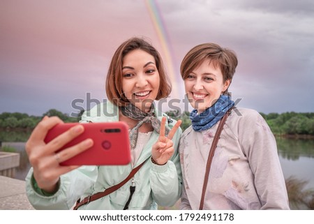 Two happy friends taking selfie photographs on mobile phones with bright rainbow at the background