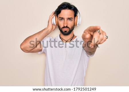 Young handsome hispanic man wearing headphones listening to music over isolated background pointing with finger to the camera and to you, hand sign, positive and confident gesture from the front