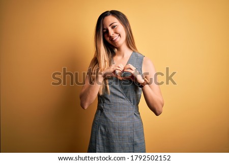 Young beautiful blonde woman with blue eyes wearing dress over yellow background smiling in love showing heart symbol and shape with hands. Romantic concept.