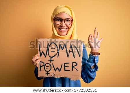 Beautiful woman with curly hair wearing muslim hijab asking for women rights holding banner doing ok sign with fingers, excellent symbol