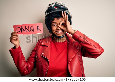 Young African American afro motorcyclist woman with curly hair holding danger paper message with happy face smiling doing ok sign with hand on eye looking through fingers