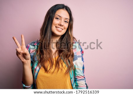 Young beautiful brunette woman wearing casual colorful shirt standing over pink background showing and pointing up with fingers number two while smiling confident and happy.