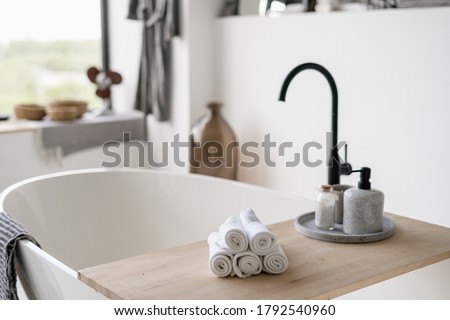 Selective focus on clean folded towels at wooden shelf on empty and contemporary tub with modern black water tap on blurred background. House decor at bright home with white interior design Royalty-Free Stock Photo #1792540960
