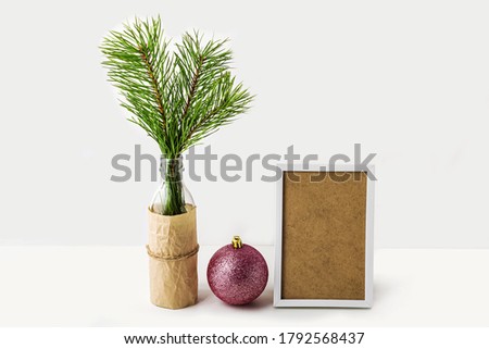 New year still life with Christmas tree branch in glass bottle,  
white frame and pink ball on white background. Mock up, copy space for text and lettering, front view