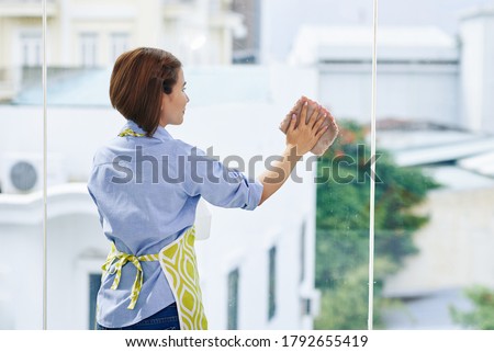 Housewife in apron cleaning big windows in her apartment Royalty-Free Stock Photo #1792655419