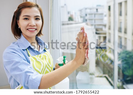 Portrait of pretty smiling Asian housewife wiping apartment window and looking at camera Royalty-Free Stock Photo #1792655422