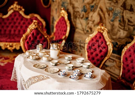Close-up of three types of tea set on a table with lace tablecloth and chairs with gold rims and red trim.