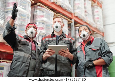 Young contemporary intercultural warehouse workers in respirators and workwear discussing new assortment of goods by huge rack