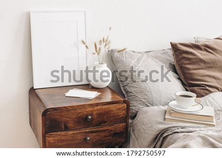 Portrait white frame mockup on retro wooden bedside table. Modern white ceramic vase, dry grass. Cup of coffee and books in bed. Beige linen pillows in bedroom. Scandinavian interior. Royalty-Free Stock Photo #1792750597