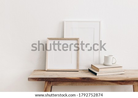 Wooden square and white portrait frame mockups on vintage bench, table. Cup of coffee on pile of books. White wall background. Scandinavian interior, neutral color palette. Artistic display  concept.