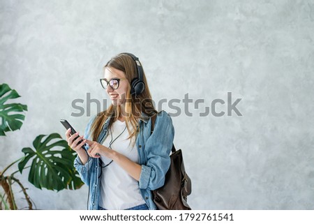 Smiling caucasian girl in eyeglasses using mobile,listening to music with headphones isolated on grey wall background indoor.Woman wearing denim clothes and brown backpack.Distance education.Lifestyle