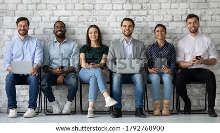 Portrait smiling diverse candidates sitting on chairs in row, looking at camera, business people applicants interns waiting for job interview, holding gadgets, employments and recruitment concept Royalty-Free Stock Photo #1792768900