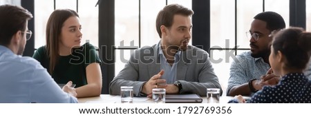 Wide image confident businessman speaking, sharing opinion, discussing project at corporate meeting with diverse colleagues, sitting at table in boardroom, leader business coach training staff