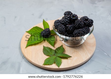 .ripe blackberries with leaves in a glass bowl on a bamboo cutting board on a concrete background, rustic, place for text