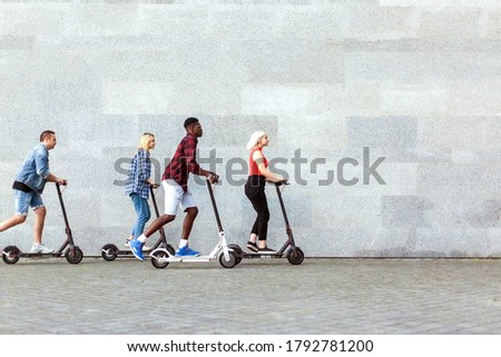 interracial company of friends rides electric scooters against the background of a wall, young people use eco transport in the city Royalty-Free Stock Photo #1792781200