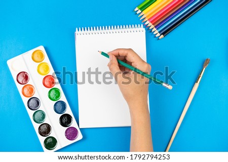 A schoolboy holds a pencil over a notebook on a blue background and school supplies. School concept. View from above.
