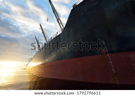 Large cargo ship (bulk carrier) anchored in port at sunset, close-up. Cranes in the background. Riga, Baltic sea, Latvia. Freight transportation, global communications, logistics, environmental damage