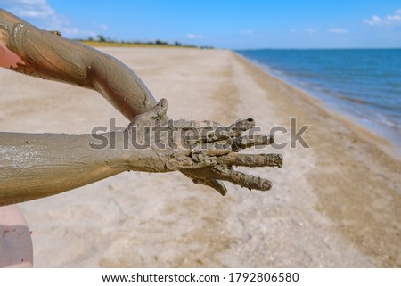 Selective focus on hands in curative mud on the background of the sea beach. The woman smears the other hand with one hand. Hot sunny day at the sea. Spa treatments. Copy space.