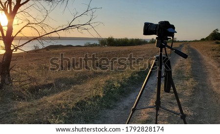 Professional photographer camera on tripod stands at dirt road near bare tree and looks to sunset sun over water. Countryside landscape with camera on stand and nobody 