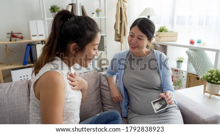 two asian japanese female friends sit on sofa and have look at ultrasonograms photo. young pregnant woman showing best sister with unborn baby in belly ultrasound picture on couch in home living room