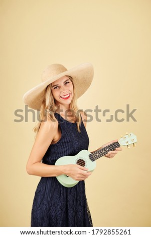 Portrait of smiling pretty blond young woman in straw hat playing ukulele, isolated on yellow