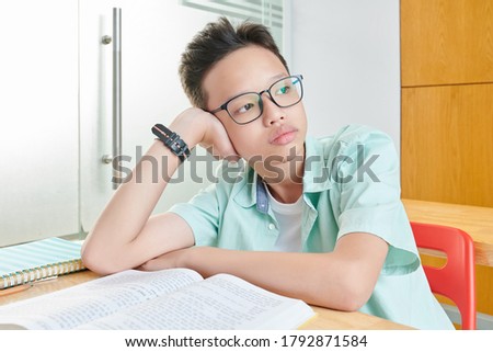 Portrait of pensive bored Vietnamese schoolboy in glasses leaning on hand and looking through window