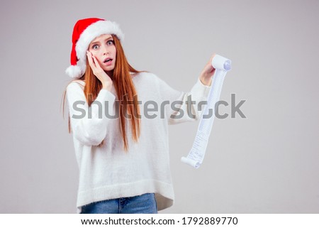 amazed and surprised redhaired ginger woman wearing red santa claus hat and knitted sweater holding long shopping receipt list in studio white background. big money xmas spending