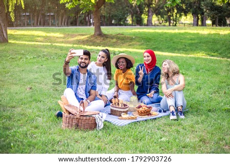 Best multiracial friends are on picnic in the park. One of the friends make selfie by smartphone.