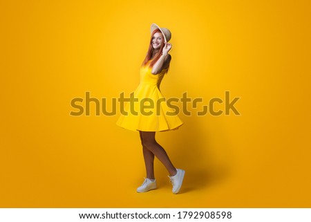 Monochrome photo of a ginger lady with hat on head and yellow dress posing on a studio wall