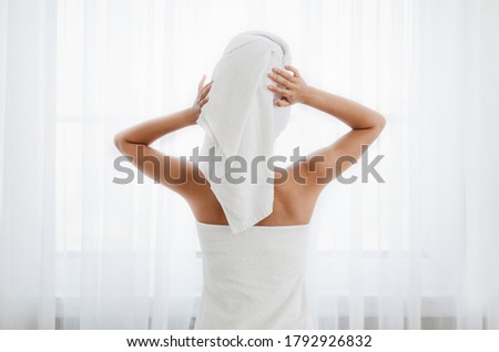 Back view of woman wrapped in towel standing next to window, drying her hair after morning shower Royalty-Free Stock Photo #1792926832