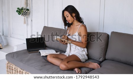 Concentrated pretty female influencer making notes of article publication for blog sitting on sofa at home interior, serious woman student writing and planning job learning online course on leisure