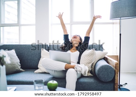 Emotional prosperous female blogger satisfied with successful project completing sitting with laptop computer, happy cheerful woman enjoying good news from social networks raising hands celebrating