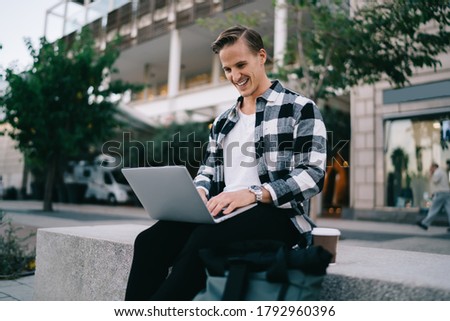 Cheerful man blogger laughing from funny photo in social networks browsing web page on laptop computer, 20s smiling caucasian hipster guy learning educational course on laptop computer in city