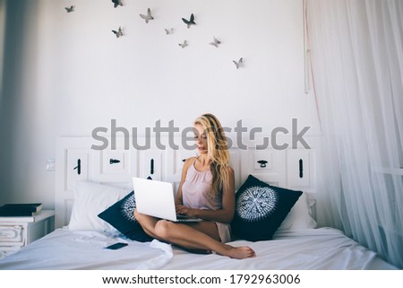 Serious blond haired female student in silk pajama searching information with laptop on comfortable bed on sunny morning in Barcelona