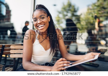 Happy smiling African American female sitting at table of summer area of modern city cafeteria while surfing Internet on tablet Royalty-Free Stock Photo #1792965877