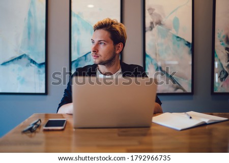 Young employee in casual clothes looking away and contemplating while sitting at desk and using laptop during work in modern workplace