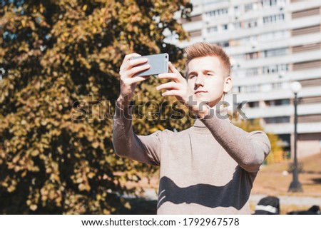 A young blond guy, dressed in a beige jacket, holds a phone in front of him and takes pictures against the background of a multi-storey building. Photo concept.