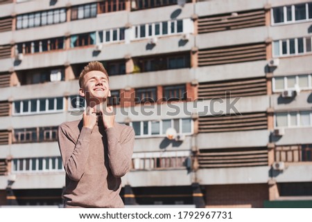A young European blond guy, dressed in a beige jacket, holds his clothes with his eyes closed against the background of a multi-storey building. Life concept.