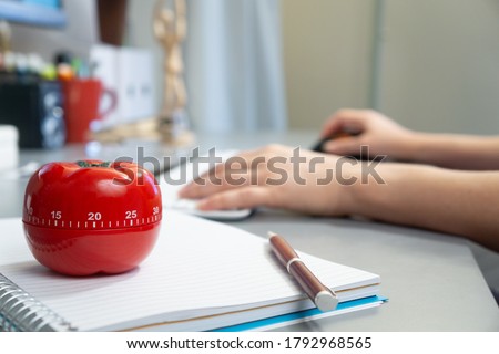 A selective focus shot of a kitchen timer in the form of a tomato on a notebook with a female working on her computer in the background Royalty-Free Stock Photo #1792968565