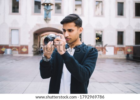 Young ethnic male photographer in smart casual outfit taking pictures on digital camera while standing on blurred background of historic building