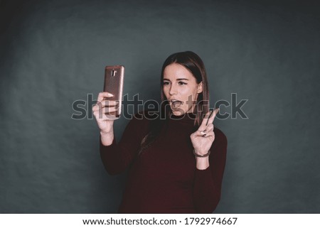 Gorgeous young female in warm brown sweater gesturing victory sign opening mouth and taking selfie on mobile phone against black background